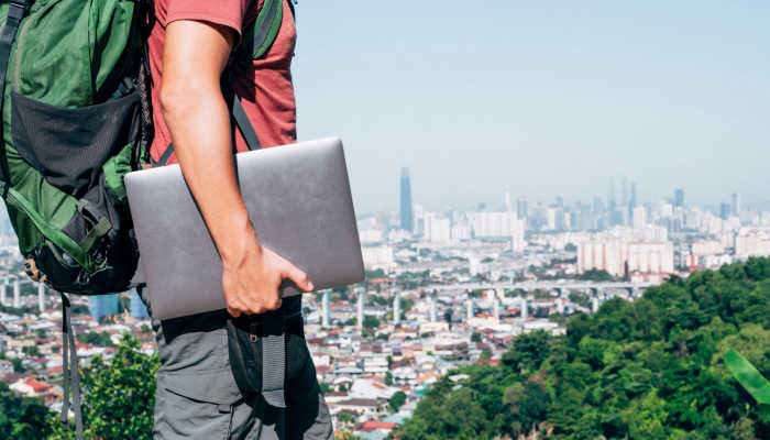Homem segurando o notebook enquanto admira uma cidade com a mochila nas costas. nywhere office e nomades digitais: como vivem as pessoas que trabalham viajando. Descubra profissões para quem quer viver dessa forma.